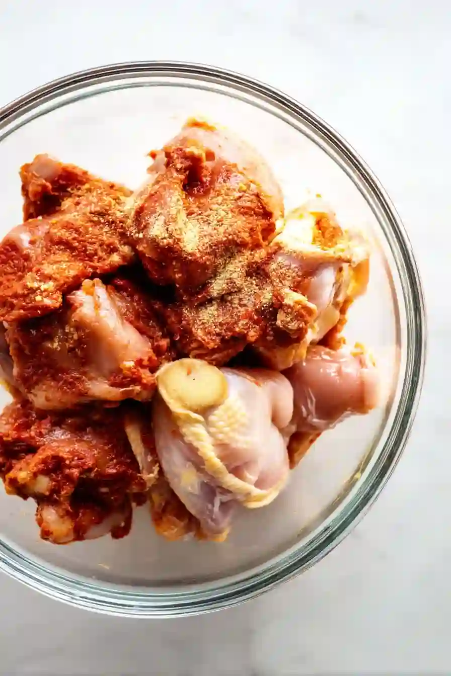 Overhead view of raw chicken pieces in a glass bowl with spices and red curry paste