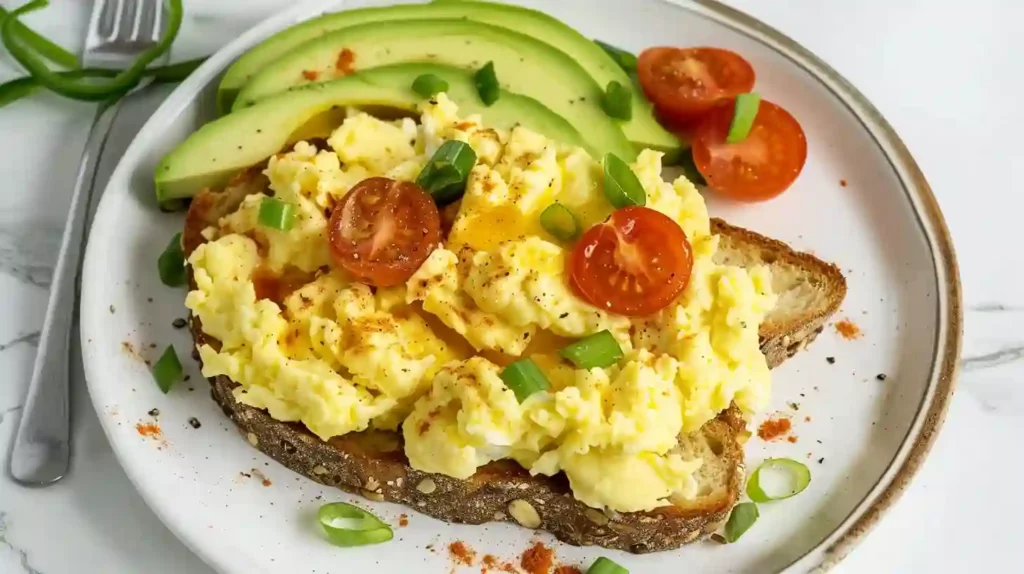 Creamy scrambled eggs served on avocado toast with cherry tomatoes on a white plate