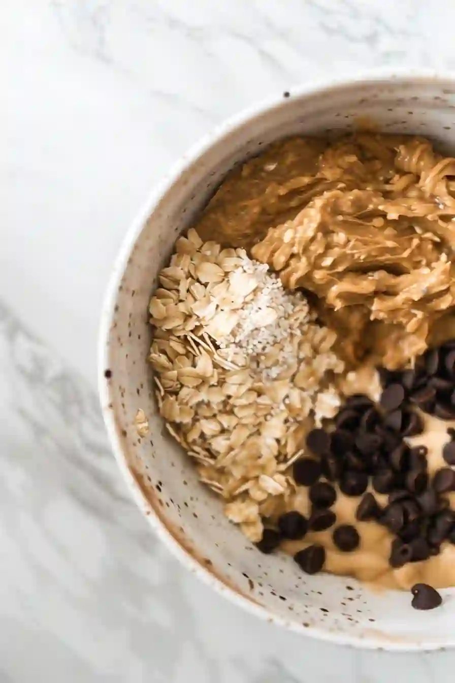 Vertical close-up of oats, peanut butter, chocolate chips, flaxseed, and honey added into a mixing bowl for peanut butter energy balls