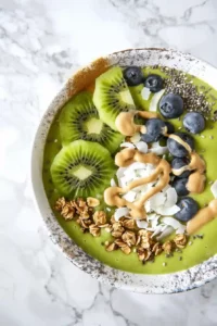 Green Protein Smoothie Bowl with Tropical Fruit 9 Green protein smoothie bowl topped with kiwi, blueberries, granola, coconut flakes, chia seeds, and peanut butter.