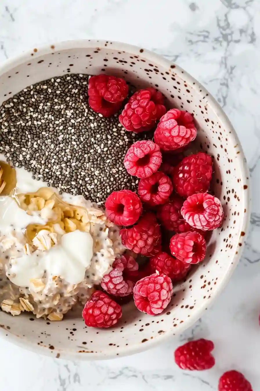 Large mixing bowl filled with oats, frozen raspberries, chia seeds, Greek yogurt, milk, vanilla extract, and honey before stirring.
